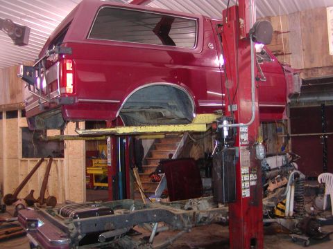 1991 Ford Bronco - The body of a red 1991 Ford Bronco is raised on a lift, undergoing disassembly in a workshop.