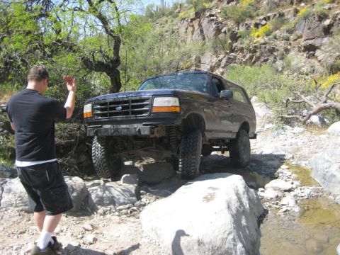 1991 Ford Bronco - A man waves as a dark blue Ford Bronco drives over rocks on a trail.