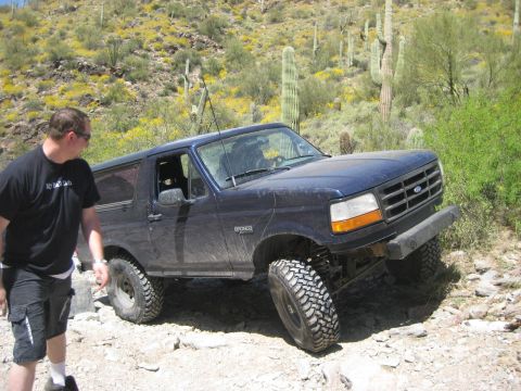 1991 Ford Bronco - A dark blue Ford Bronco navigates a rocky desert trail with saguaro cacti in the background.