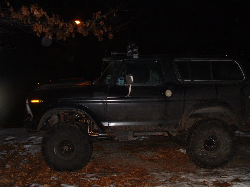 Lifted black 1970s Ford Bronco at night on snowy leaves with roof lights and mud tires