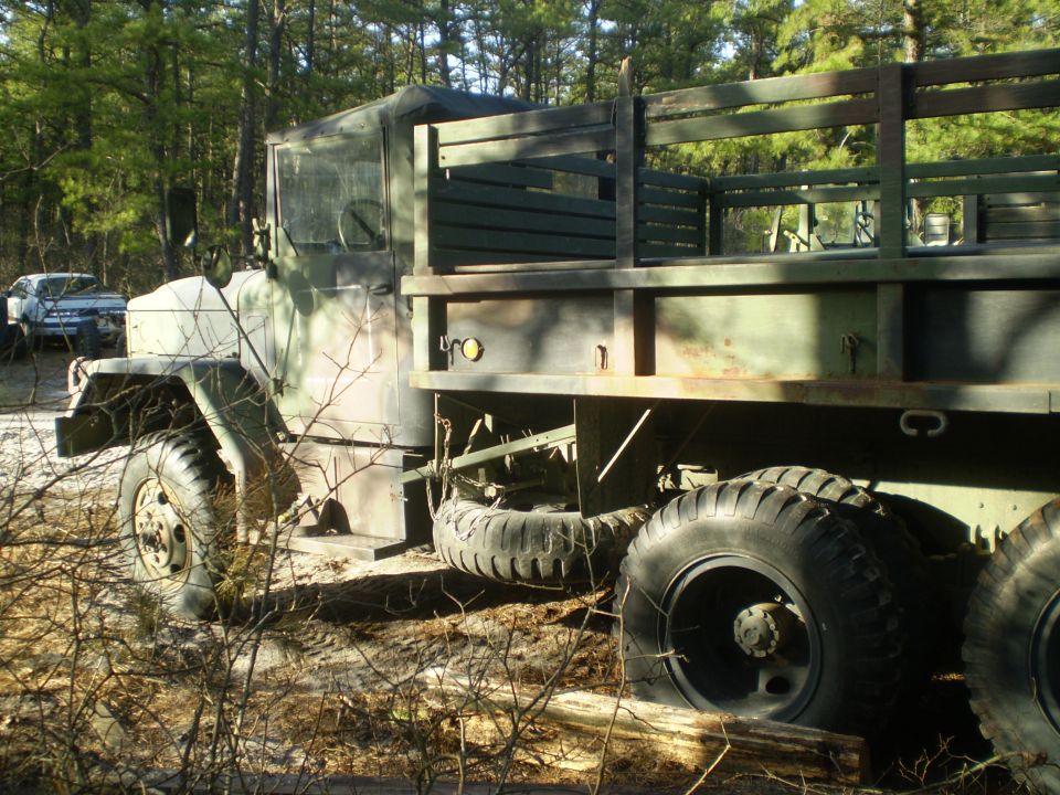Olive drab 6x6 military stake-bed truck parked in a sandy pine forest clearing