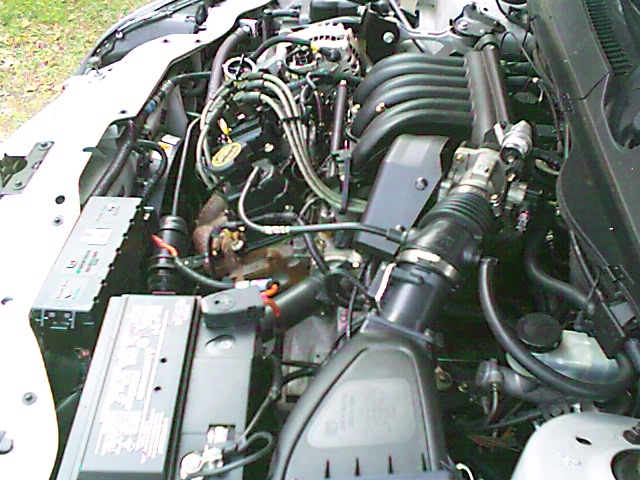Engine bay of a white car showing V6 engine, air intake tube, battery, and ignition wiring