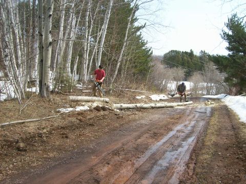 2000 Chevrolet Tahoe - Two men use chainsaws to cut fallen logs on a muddy road surrounded by trees and snow.