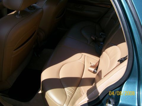 1995 Mercury Grand Marquis - Rear seat of a green sedan photographed through the open rear door in bright sun, showing the tan leather bench with sculpted seatback, bro…