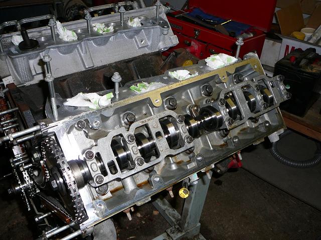 Engine block with camshafts and timing chain on a stand, with tools and a battery in the background