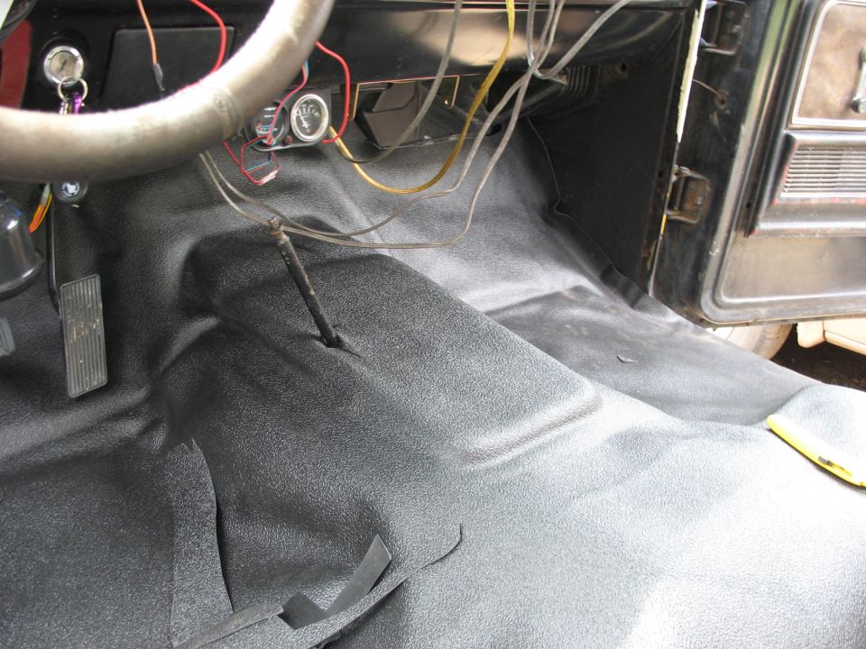 Vehicle interior floor with new black textured matting installed. Steering wheel and dashboard wiring visible