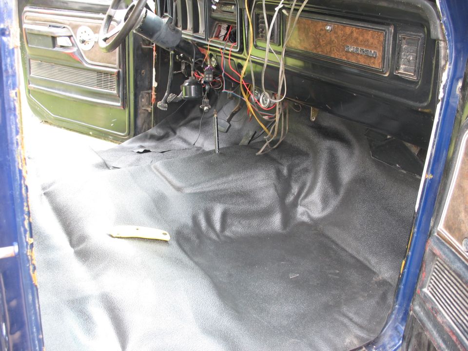 interior shot of a blue vehicle, showing dashboard, steering wheel, and floor with new black matting. Wires are exposed near the steering column