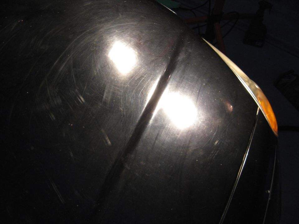 Black car hood with numerous scratches and two bright reflections. Orange turn signal visible on the right