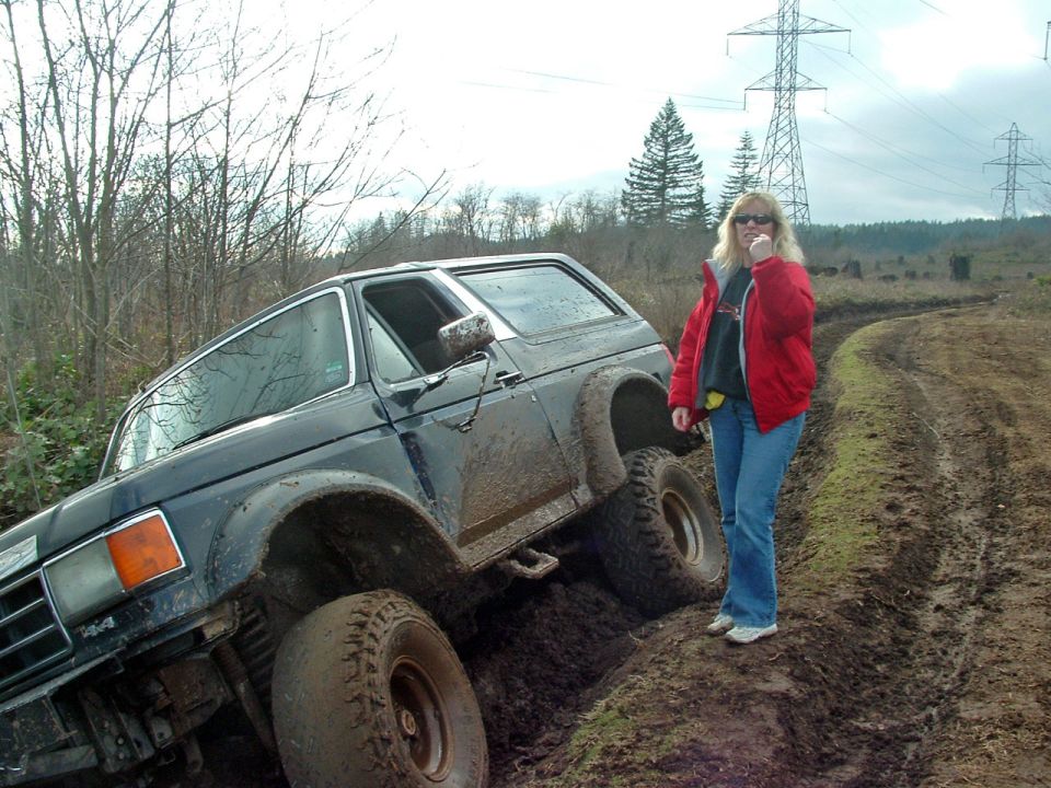 Lifted dark blue Ford Bronco stuck nose-down in mud beside a trail, woman in red jacket standing next to it