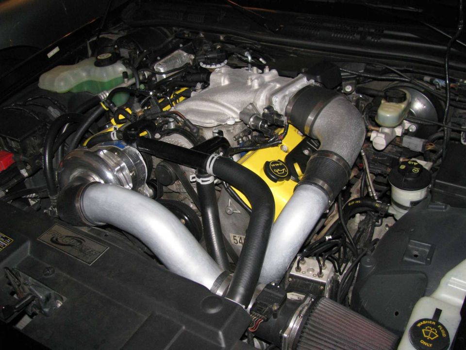Engine bay with silver turbocharger, yellow valve covers, and custom intake piping. Some hoses and wiring are visible