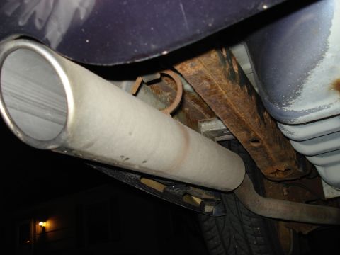 2000 Ford Crown Victoria - Close-up underside night shot of a large-diameter stainless exhaust tailpipe exiting behind the rear bumper of a dark blue sedan, shot from…