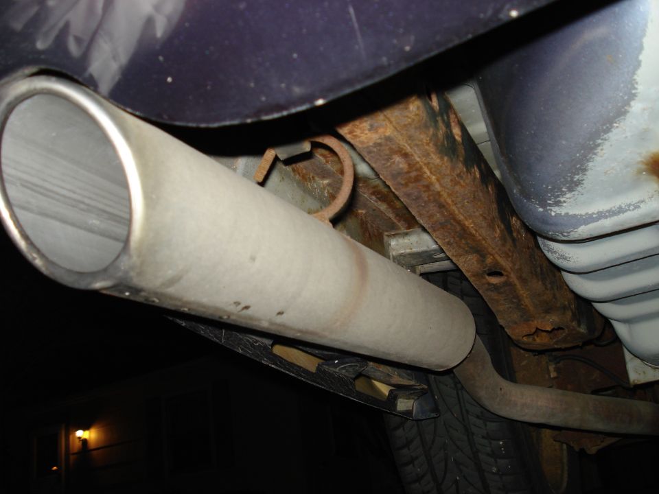 Close-up of stainless exhaust tailpipe under rear of dark blue sedan with rusty frame rail