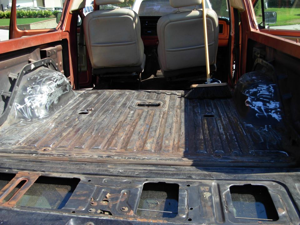 Orange 1978 Ford Bronco interior cargo area with worn metal floor and tan seats. A broom leans against the driver's seat