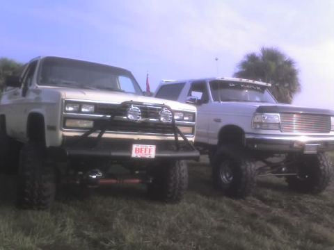 1995 Ford Bronco - A white Ford Bronco and a beige truck are parked on grass, both modified with lift kits and large tires.