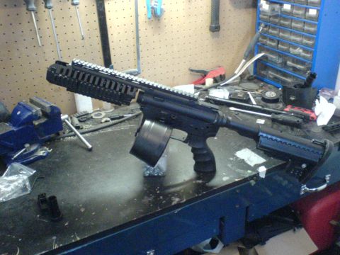 1985 Ford Bronco - An assault rifle with a drum magazine is displayed on a workbench, surrounded by tools and parts.
