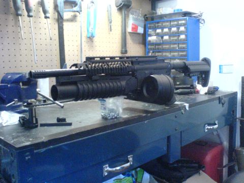 1985 Ford Bronco - A modified assault rifle with a drum magazine and grenade launcher is displayed on a workbench.