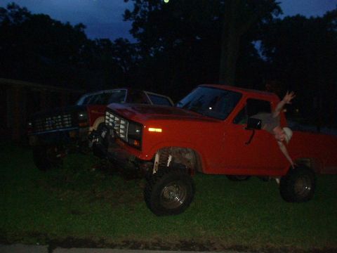 1983 Ford Bronco - A red pickup truck and a dark SUV are parked on a grassy area at dusk.