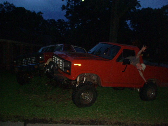 Red pickup truck with lifted suspension and a person waving from the driver's window, parked next to a dark SUV