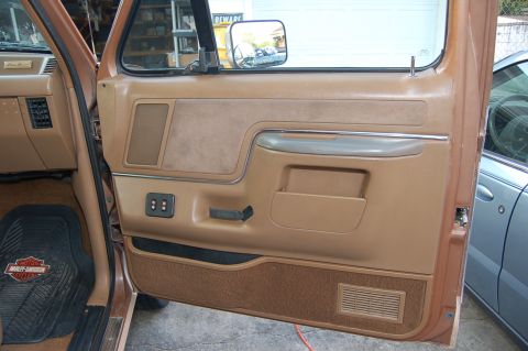 1989 Ford Bronco - The interior door panel of a brown vehicle, with a Harley Davidson floor mat and a glimpse of another car outside.