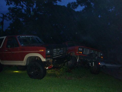 1983 Ford Bronco - Two red SUVs are parked on a grassy area at dusk. The front vehicle features a black hood and appears to be a Ford Bronco.