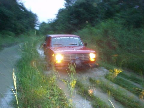 1983 Ford Bronco - This red Ford Bronco is navigating a challenging muddy path, surrounded by lush green vegetation.