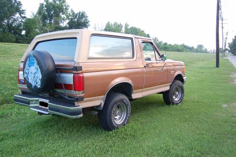 1989 Ford Bronco - A brown Ford Bronco is parked on a grassy area next to a road with trees in the background.