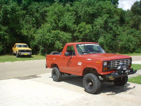 1983 Ford Bronco - A red Ford Bronco is parked on a driveway, with a yellow Ford pickup truck in the background.
