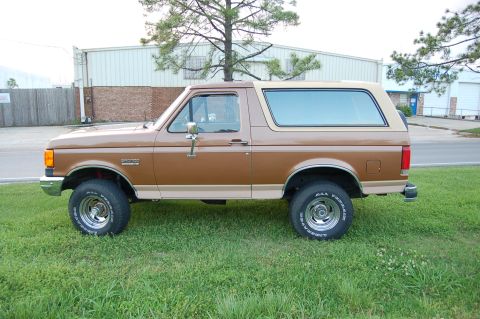 1989 Ford Bronco - This two-tone brown Ford Bronco is shown from the side, parked on a grassy area with a building in the background.