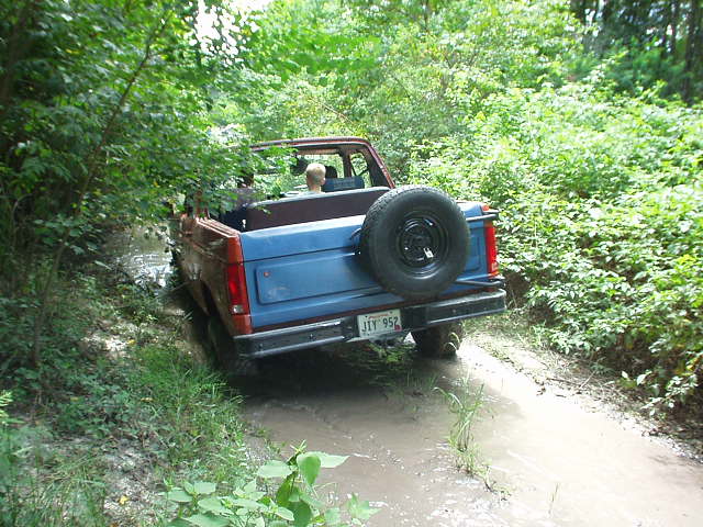 Rear view of a blue and red 1983 Ford Bronco driving through mud on a wooded trail