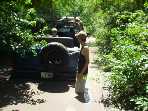 1983 Ford Bronco - Two off-road vehicles navigate a muddy trail surrounded by dense foliage.