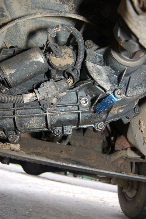 1989 Ford Bronco - Close-up of the dirty drivetrain components, including the transmission and transfer case, with a piece of blue tape attached.