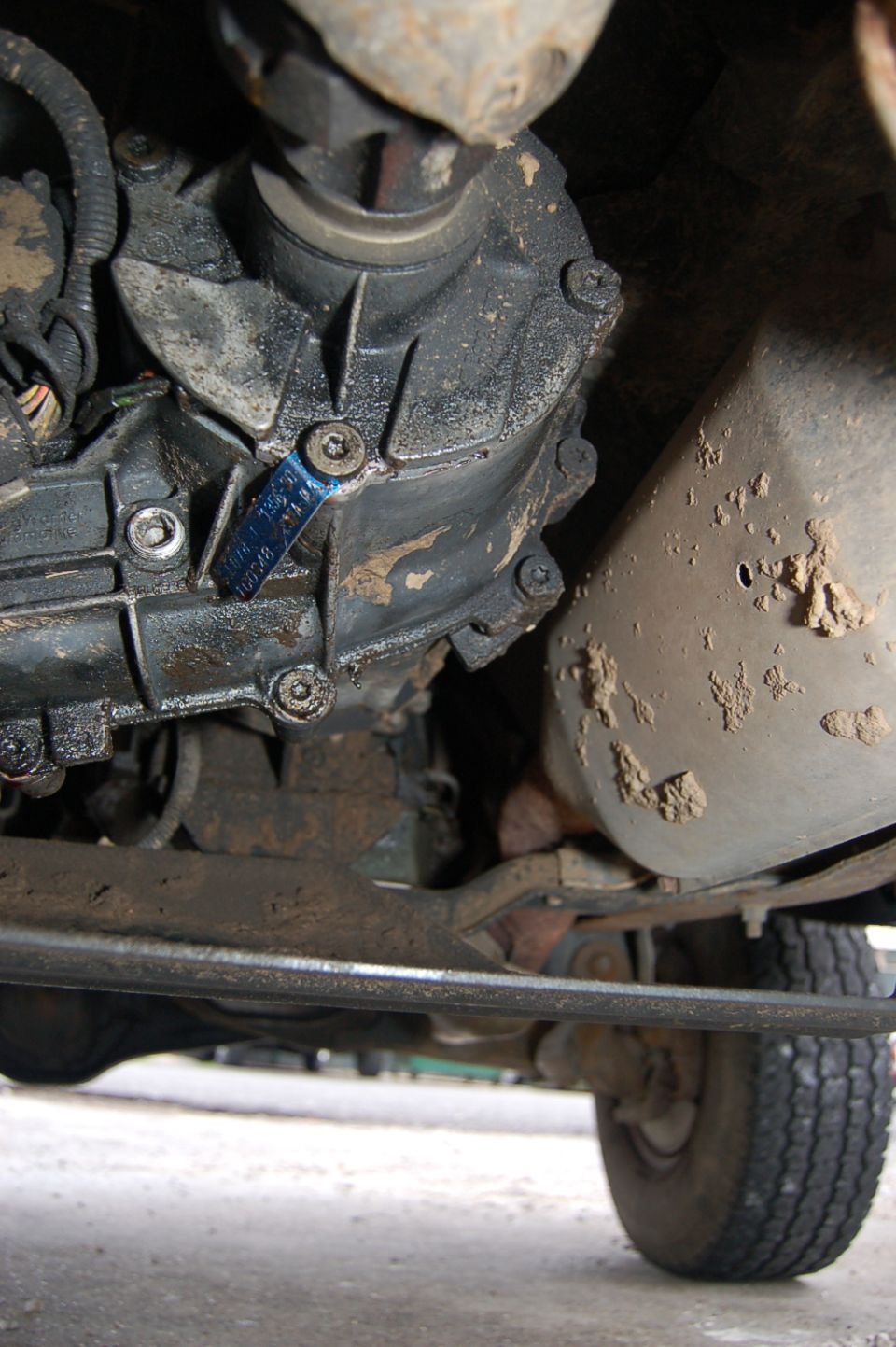 Underside view of a dirty drivetrain with a blue tag on the transfer case and a portion of the exhaust pipe