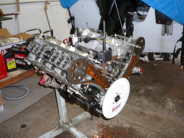 Engine on a stand with timing gears and chains visible. A white disc with red lettering is attached to the front