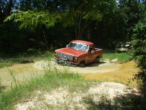 1983 Ford Bronco - A red pickup truck navigates a muddy trail, with another vehicle visible in the background.