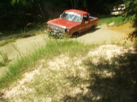 1983 Ford Bronco - A red pickup truck is stuck in the mud on a dirt path, with another vehicle in the distance.