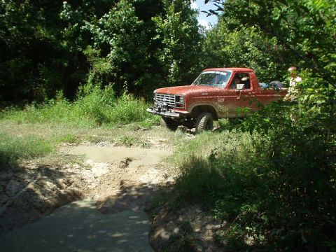 1983 Ford Bronco - A red pickup truck navigates a muddy trail surrounded by dense green foliage.