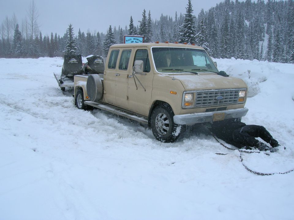Tan van with a trailer carrying two snowmobiles, stuck in snow. A person is underneath the front