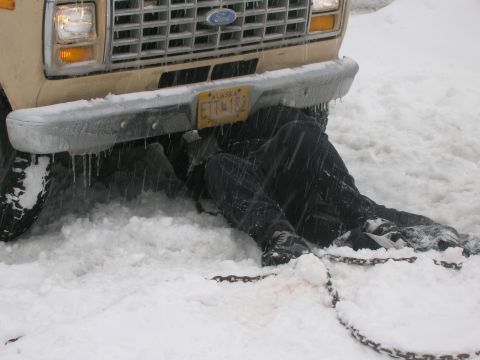 2003 Jeep TJ - A person works on a Ford van stuck in the snow, with chains visible on the ground.