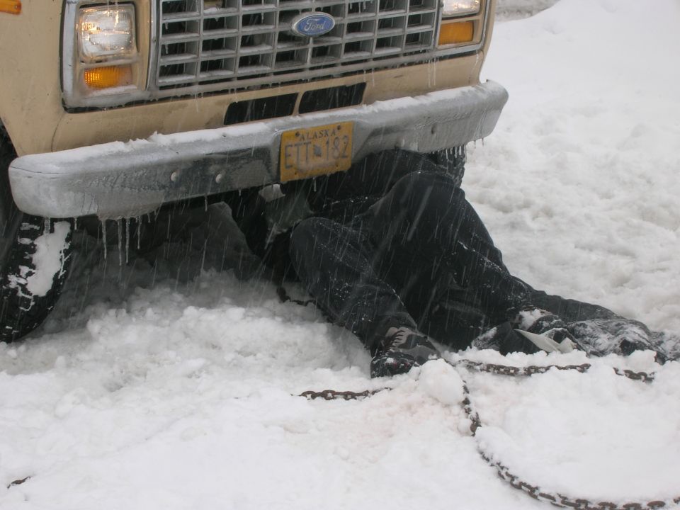 Ford van front end with person underneath in snow, Alaska license plate ETT-182