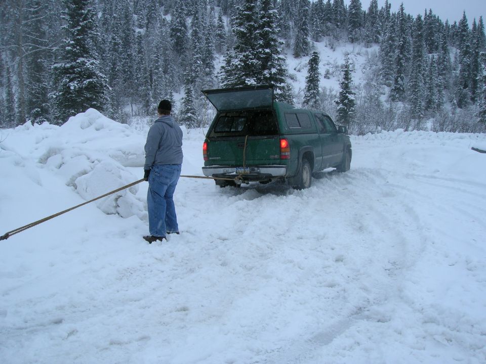 Green pickup truck with cap, rear view, in snowy area. Person holding a long pole near truck