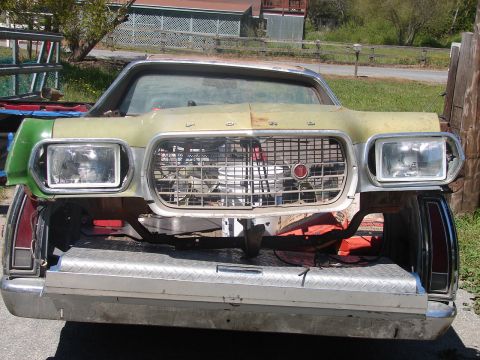 1973 Ford Ranchero - Head-on view of a faded green 1970s Ford's front clip with the bumper removed, showing the chrome egg-crate grille with red-and-white cente…