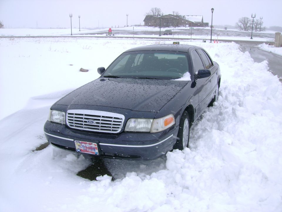 Dark sedan partially buried in snowdrift, front view. Grille suggests a 1998 model