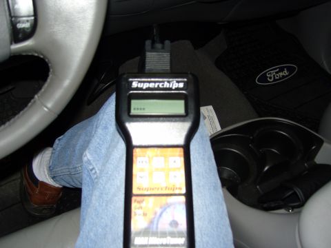 2007 Ford Mustang - A Superchips tuner device is held in a vehicle's interior, with a Ford floor mat in the background.