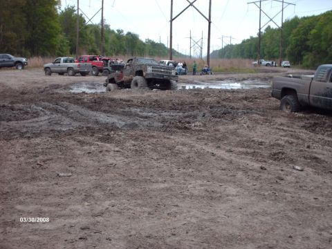 1982 Ford F-100 - Several trucks are stuck in a muddy area, with some people watching. Power lines are visible in the background.