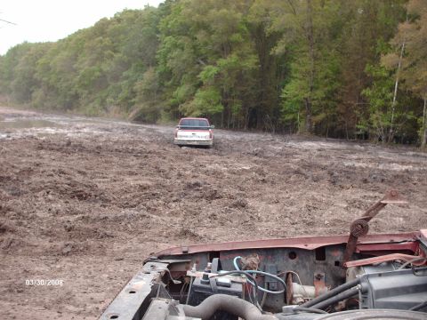 1982 Ford F-100 - A Chevrolet pickup truck is stuck in a muddy field with trees in the background. The foreground shows the engine of another vehicle.