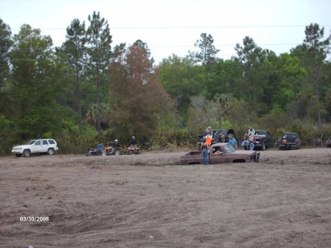 1982 Ford F-100 - A group of people gather around a rusty pickup truck that is stuck in the mud at an outdoor event.