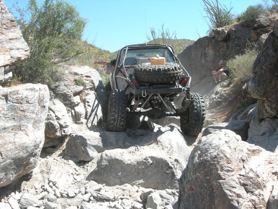 Off-road vehicle with spare tire and jerry can on rear, climbing over large rocks on a narrow trail