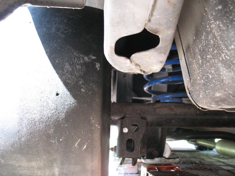 Close-up of a blue coil spring and suspension parts under a vehicle. A Supermotors.net logo is visible