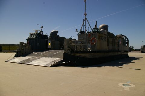 1986 Ford Bronco - Two military hovercraft, identified as LCACs, are positioned on a paved area with a bright blue sky overhead.