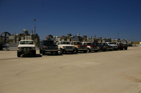 1986 Ford Bronco - Several Ford trucks and SUVs are parked in a line with hovercraft and military vessels behind them.
